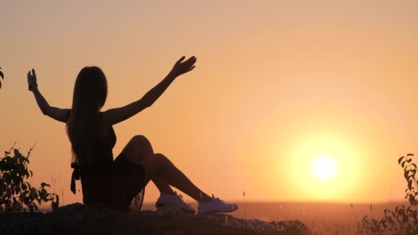 Joven mujer elegante en vestido corto negro sentado en una roca relajante al aire libre en la noche de verano. Mujer de moda disfrutando de la cálida puesta de sol en la naturaleza. - Metraje, vídeo