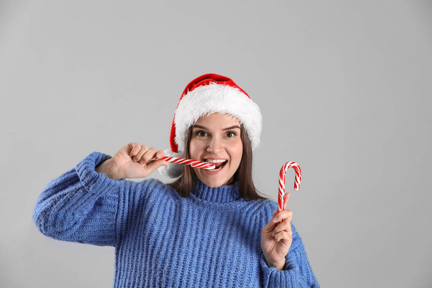 Pretty woman in Santa hat and blue sweater eating candy cane on grey background - Фото, изображение