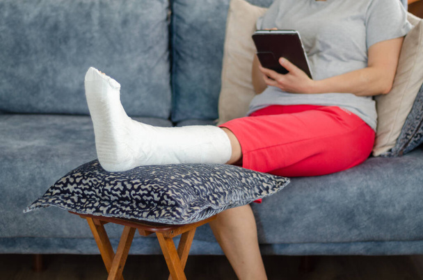 Женщина средних лет со сломанной ногой сидит на диване и читает сообщения дома. - Фото, изображение