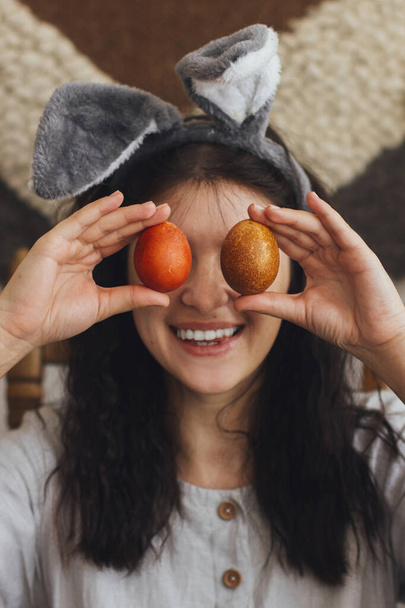 Szczęśliwa młoda kobieta w króliczych uszach i sukni lnianej trzymając pisanki w oczach i uśmiechając się na tle rustykalnym pokoju. Szczęśliwej Wielkanocy! Naturalne barwione jaja wielkanocne w rękach, zabawne chwile - Zdjęcie, obraz