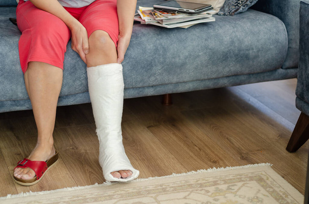 Женщина средних лет со сломанной ногой сидит на диване и занимается спортом. - Фото, изображение