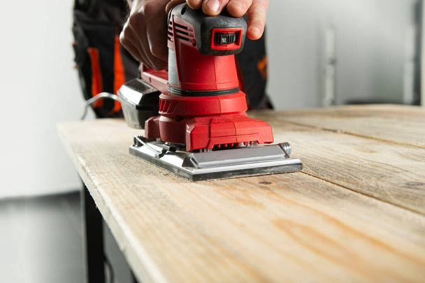 Elektrischer Bandschleifer, Schleifmaschine in Männerhand. Bearbeitung des Werkstücks auf hellbraunem Holztisch  - Foto, Bild