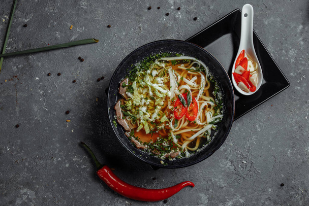 Cucina asiatica, zuppa di manzo in un piatto nero su sfondo scuro. Zuppa Fo con brodo di manzo piccante, filetto di manzo, tagliatelle udon, pasta di peperoncino - Foto, immagini