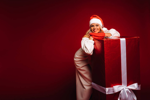 portrait of a smiling girl in a white dress and Santa hat with a huge Christmas gift on a red background - Zdjęcie, obraz