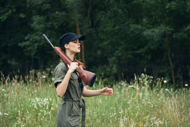 Woman on nature the gun on the shoulder green jumpsuit black cap green leaves  - Photo, image