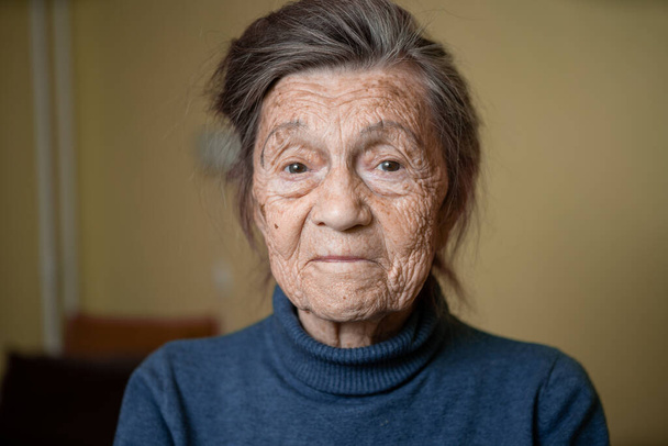 90-vuotias söpö vanha nainen, jolla on harmaat hiukset ja ryppyjä kasvot, yllään villapaita, muotokuva suuri, hymyilevä ja näyttää iloisesti, huoneen tausta. Teema Pitkäaikainen maksa ja ikääntyminen, vanhukset hyvällä tuulella. - Valokuva, kuva