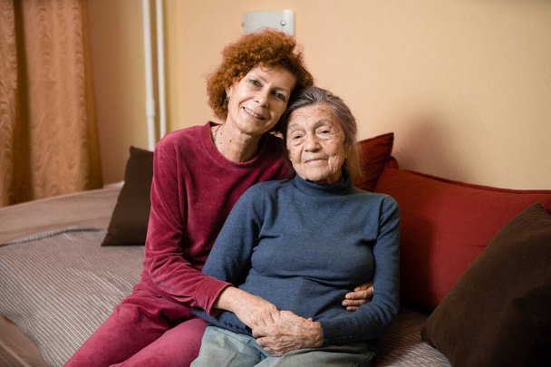 Eine ältere kaukasische Frau umarmt zärtlich ihre neunzigjährige Mutter mit grauen Haaren, Gesicht mit tiefen Falten, zu Hause auf dem Sofa, lächelnd Mutter und Tochter. Thema Pflege und Engagement für alte Eltern. - Foto, Bild