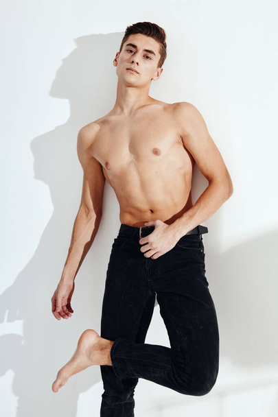 A half-naked guy in black pants stands on one leg with his elbows on the wall - Foto, Imagen