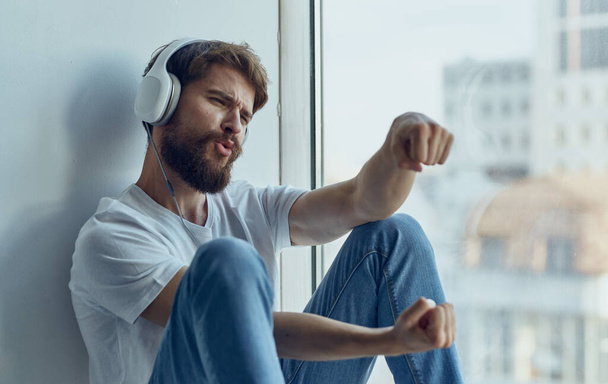 een man met een koptelefoon zit op de vensterbank en luistert naar muziek imitatie besturen van een auto stuurwiel Studio - Foto, afbeelding