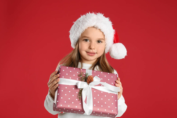Cute child in Santa hat with Christmas gift on red background - Zdjęcie, obraz