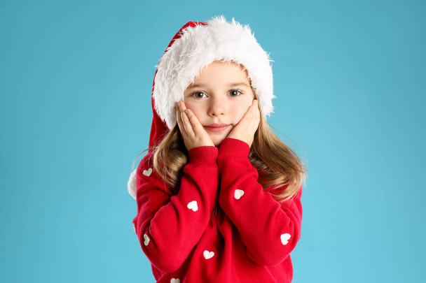 Cute child in Santa hat on light blue background. Christmas celebration - Fotó, kép