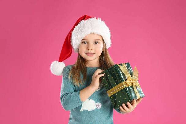 Cute child in Santa hat with Christmas gift on pink background - Photo, image