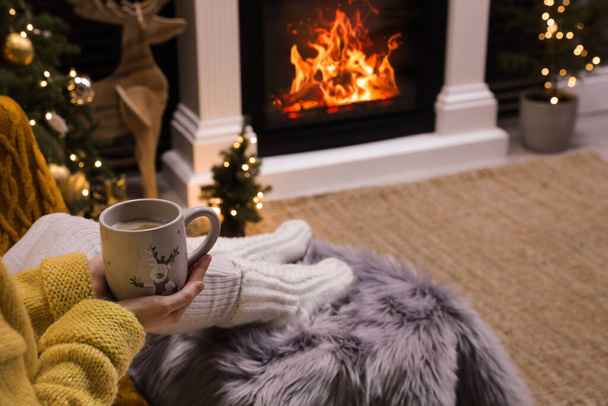 Woman with cup of drink sitting near burning fireplace at home, closeup - Photo, image