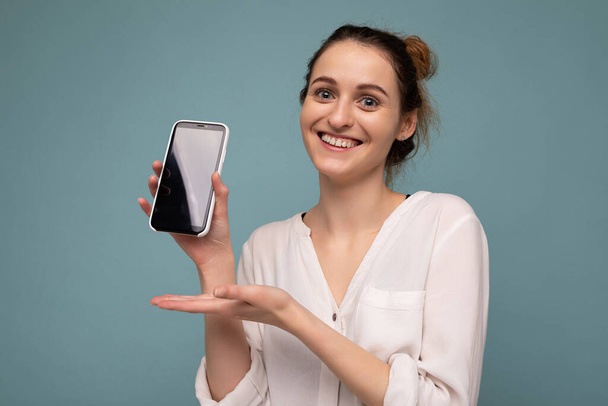 Beautiful positive smiling young blonde curly woman wearing casual white shirt and isolated over blue background wall holding phone and showing smartphone with empty dcreen for mockup looking at - Zdjęcie, obraz