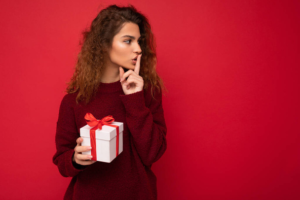 Photo de belle jeune femme bouclée brune positive isolée sur un mur de fond rouge portant un pull rouge tenant une boîte-cadeau regardant sur le côté et montrant un geste shh - Photo, image