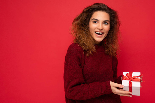 Приваблива позитивна здивована молода брюнетка кучерява жінка ізольована на червоному тлі стіни в червоному светрі тримає подарункову коробку, дивлячись на камеру
 - Фото, зображення