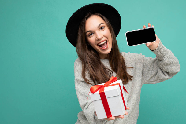 Photo of beautiful happy joyful young brunette woman isolated over blue background wall wearing stylish black hat and grey sweater holding gift box showing mobile phone screen display for mockup and - Photo, image