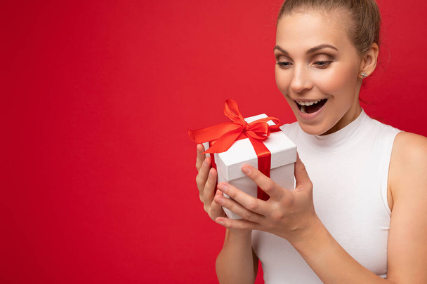 Photo shot of pretty positive surprised young blond woman isolated over colourful background wall wearing trendy outfit look holding gift box and looking at present box with red ribbon - Φωτογραφία, εικόνα