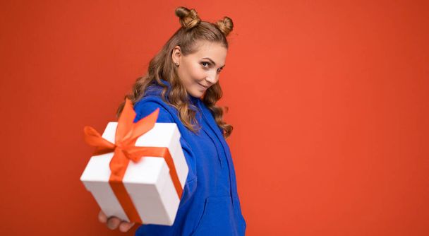 Shot of attractive happy young blonde woman isolated over red background wall wearing blue trendy hoodie holding gift box and looking at camera - Фото, зображення