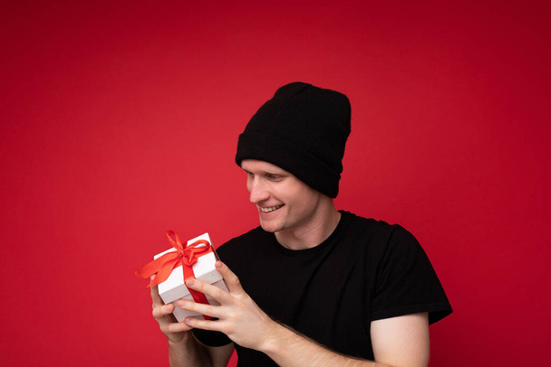 Hezký šťastný usmívající se mladý muž izolované přes červené pozadí zeď nosí černý klobouk a černé tričko drží bílou dárkovou krabici s červenou stuhou a při pohledu na současnost - Fotografie, Obrázek