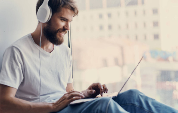 ευτυχισμένος άνθρωπος με ακουστικά κάθεται στο περβάζι του παραθύρου και κοιτάζοντας οθόνη laptop - Φωτογραφία, εικόνα