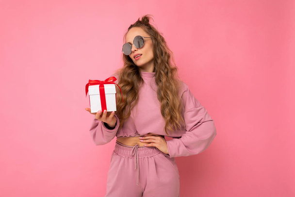 Beautiful happy young brunette woman isolated over colourful background wall wearing stylish casual clothes holding gift box and looking to the side - Foto, Imagem