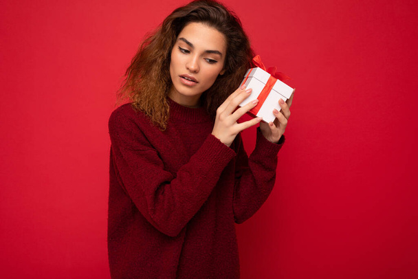Atraktivní pozitivní koncentrované mladé brunet kudrnaté žena izolované přes červené pozadí zeď na sobě červený svetr držení dárkové krabice a snaží se hádat překvapení - Fotografie, Obrázek
