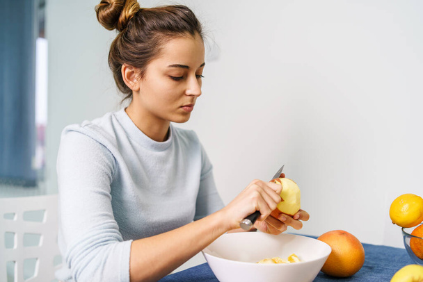 Jeune femme caucasienne épluchant des fruits assis à la table à la maison - vue de côté sur une jolie fille tenant un couteau et préparant des pommes - manger sainement concept copier l'espace - Photo, image