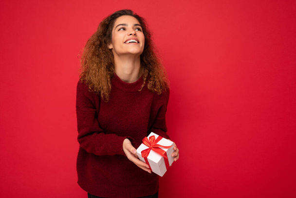 Привлекательный позитивный радостный юная брюнетка кудрявая женщина человек изолирован на красном фоне стены в красном свитере держа подарочную коробку глядя вверх - Фото, изображение