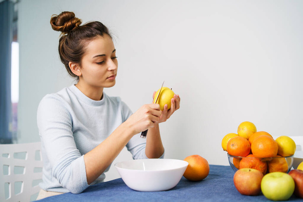 Mujer joven caucásica pelando fruta mientras está sentada junto a la mesa en casa - vista lateral de la niña bonita sosteniendo el cuchillo y la manzana preparando la comida - concepto de alimentación saludable copiar espacio - Foto, Imagen