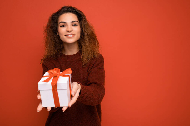 Gyönyörű boldog fiatal barna nő elszigetelt át színes háttér fal visel elegáns alkalmi ruhák gazdaság ajándék doboz és nézi a kamera - Fotó, kép