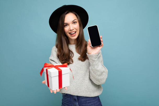 Знімок досить усміхненої позитивної молодої брюнетки, ізольованої на синьому фоні стіни в стильному чорному капелюсі і сірому светрі, що тримає подарункову коробку, показуючи екран смартфона для маскування і
 - Фото, зображення