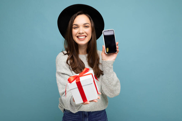 Shot of pretty smiling positive young brunet woman isolated on blue background wall wearing black hat and grey sweater holding gift box showing smartphone screen display for mockup and looking at - Fotografie, Obrázek