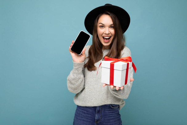 Захоплююча щаслива молода брюнетка ізольована на синій стіні в стильному чорному капелюсі і сірому светрі, що тримає подарункову коробку, показуючи мобільний телефон для маскування і дивлячись на
 - Фото, зображення