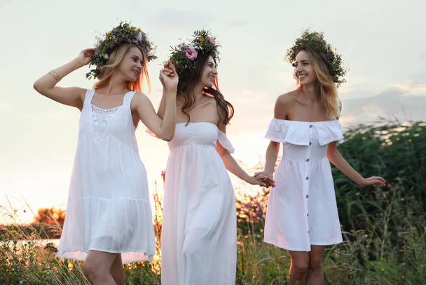 Young women wearing wreaths made of beautiful flowers outdoors at sunset - Фото, зображення