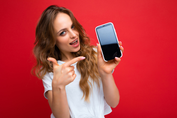 Photo of beautiful smiling young woman good looking wearing casual stylish outfit standing isolated on background with copy space holding smartphone showing phone in hand with empty screen display for - Фото, изображение