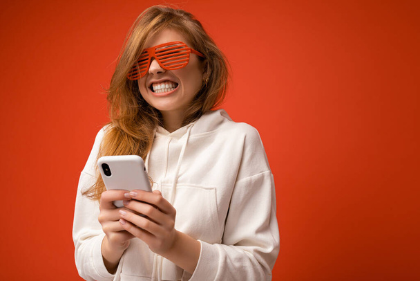 Piękne emocjonalne młoda blondynka kobieta w stylowej białej bluzie i śmieszne kolorowe okulary stojąc w izolacji nad kolorowe tło gospodarstwa i za pomocą telefonu komórkowego komunikacji online na - Zdjęcie, obraz