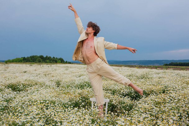 Tall handsome man dressed in a white suit on naked body dancing on the white chair in camomile flowers field - Foto, Bild