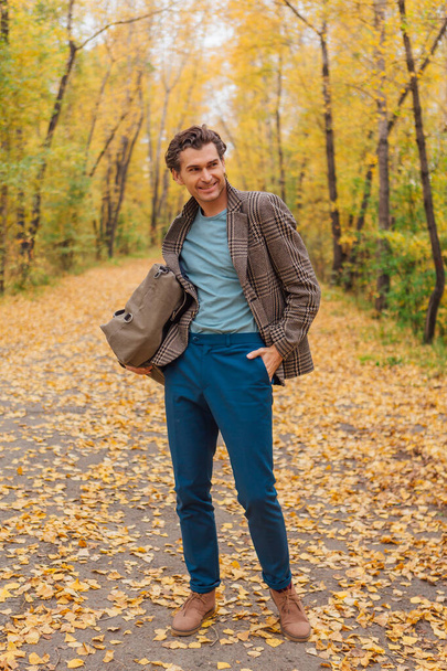 Tall handsome man dressed in a brown jacket walking with a bag on the autumn alley - Foto, immagini