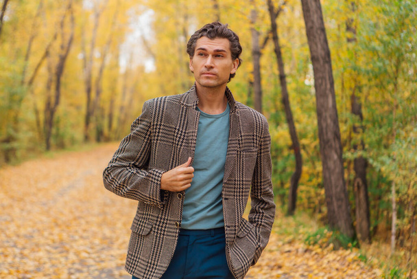 Tall handsome man dressed in a brown jacket walking on the autumn alley - Photo, image