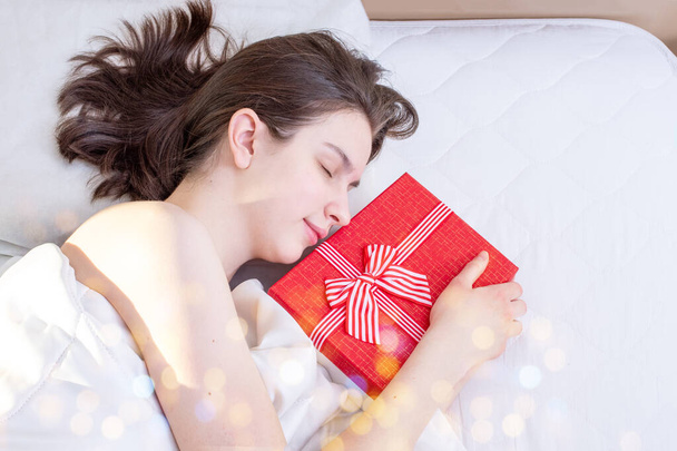 Mattina di compleanno, San Valentino, giorno del matrimonio, Natale. Giovane ragazza bruna addormentata con scatola regalo rossa sul letto a casa, vista dall'alto, copia spazio, bokeh - Foto, immagini
