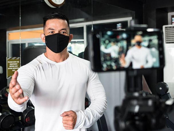 Beyaz kazak giyen kaslı Asyalı bir adam ve siyah yüz maskesi bir fitness vlogu kaydediyor. Sahne arkasında. Spor salonunda.. - Fotoğraf, Görsel