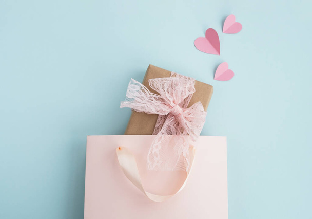 Valentijnsdag en vakantie concept. Cadeaudoosje in boodschappentas en roze hartjes op pastelblauwe achtergrond. Minimale samenstelling. Bovenaanzicht, flat lay, kopieerruimte - Foto, afbeelding
