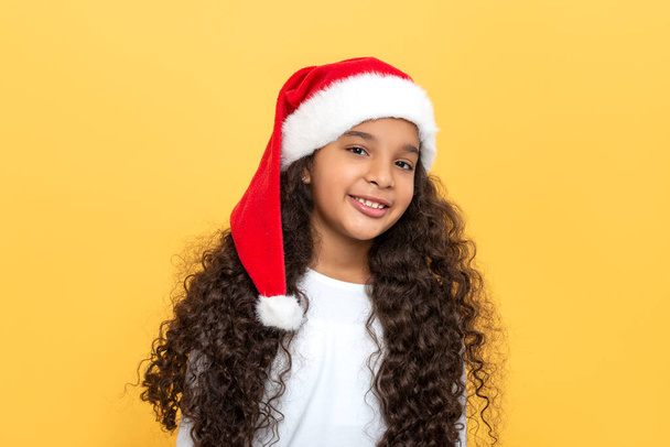 Tailliertes Porträt eines dunkelhäutigen jungen Mädchens mit langen lockigen Haaren in Weihnachtsmannmütze auf gelbem Hintergrund - Foto, Bild