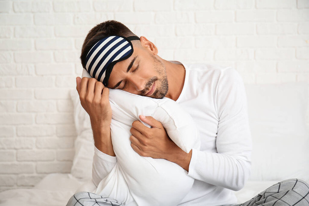 Portrait of young man with sleep mask and pillow on bed in bedroom - Photo, Image