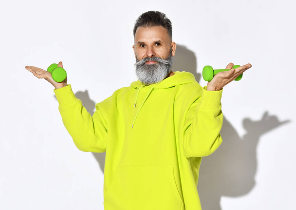 Portrait of happy active old brutal bearded man in yellow hoodie holding green dumbbells in hands on open palms - Fotó, kép