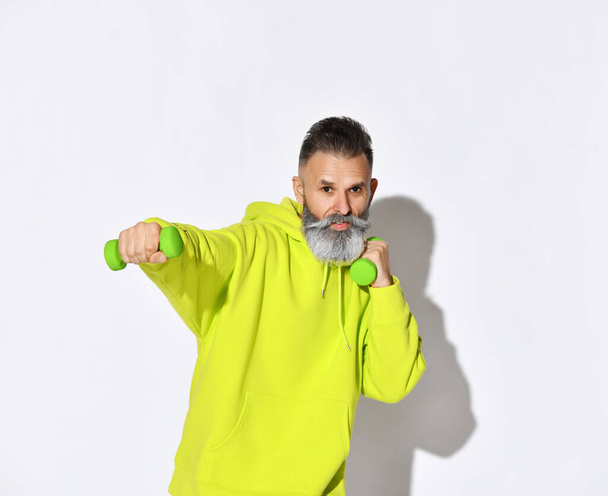 Happy active old brutal bearded man in yellow hoodie enjoys fitness training boxing with green dumbbells in hands - Fotó, kép