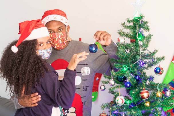 Linda pareja joven con máscaras protectoras cerca de un árbol de Navidad. Diferentes Navidad en el concepto de bloqueo. Navidad, coronavirus y concepto de amor. - Foto, imagen