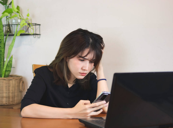 Portrait of upset Asian woman wearing black shirt, sitting at wooden table with computer laptop, reading message in mobile phone, touching head, feeling worried. white wall with house plant background. - Photo, Image