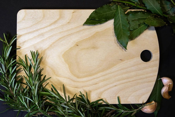 Cutting board of light wood framed with bay leaves, rosemary sprigs and cloves of garlic, designer template  - Photo, Image